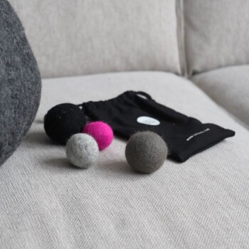 Eine schwarze Zugtasche mit der Aufschrift "Lucy Balu" enthält drei bunte Filzkugeln in Grau, Grün und Schwarz, wobei eine Kugel den Markennamen trägt.