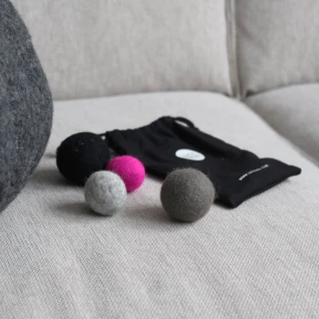 Eine schwarze Zugtasche mit der Aufschrift "Lucy Balu" enthält drei bunte Filzkugeln in Grau, Grün und Schwarz, wobei eine Kugel den Markennamen trägt.
