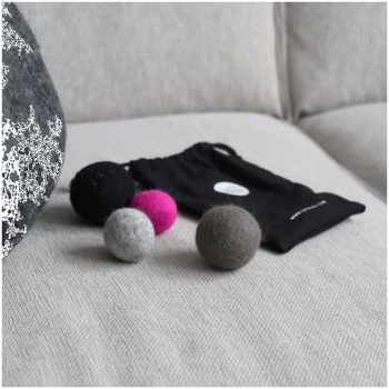 Eine schwarze Zugtasche mit der Aufschrift "Lucy Balu" enthält drei bunte Filzkugeln in Grau, Grün und Schwarz, wobei eine Kugel den Markennamen trägt.