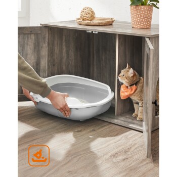 Eine Person stellt eine graue Katzenstreuschachtel auf einen Holzboden, während eine neugierige Katze mit einem orangefarbenen Halstuch von einem nahegelegenen Schrank zusieht.