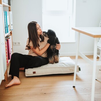 Frau sitzt auf dem Boden und kuschelt mit einem kleinen schwarzen lockigen Hund auf einem beigen Haustierbett am Fenster in einem hellen Raum; kein lesbarer Text.