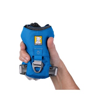 Eine Person hält ein blaues Hundegeschirr mit einem Ruffwear-Logo, das einen Metall-D-Ring und verstellbare Riemen hat.