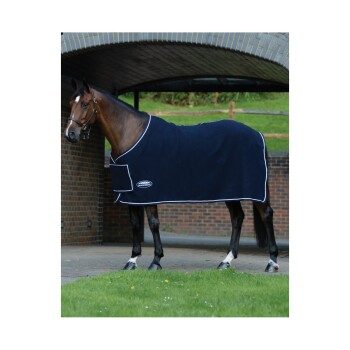 Ein braunes Pferd in einer marineblauen Fleece-Decke mit weißem Rand steht in einem grasbewachsenen Bereich nahe einer Ziegelmauer und einem Torbogen.