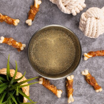 Metall-Hundenapf gefüllt mit Sand, umgeben von Hundekauknochen und zwei weißen Seilspielzeugen auf grauer Oberfläche.