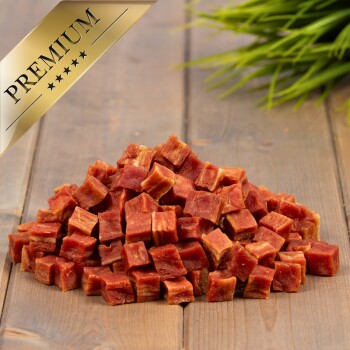 Ein Haufen kleiner, gewürfelter, roter Haustier-Leckerlis auf einer Holzoberfläche, mit einem goldenen Etikett "PREMIUM" in der oberen linken Ecke.