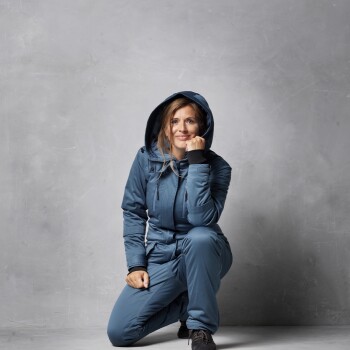 Eine Person in einer blauen Kapuzenjacke und passenden Hosen kniet vor einem grauen Hintergrund und zeigt einen lässigen Outdoor-Stil.