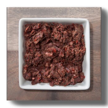 Ein quadratisches weißes Geschirr gefüllt mit einer groben, rohen Tiernahrungsmischung, die dunkle rote und braune Farbtöne aufweist, auf einer Holzoberfläche platziert.