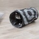 Un chat noir et blanc regarde à travers un jouet tunnel rayé gris et noir, posé sur un sol en bois.