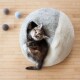 Ein gemütliches, rundes grau-creamfarbenes Katzenbett mit einer schlafenden Katze darin, umgeben von drei bunten Filzkugeln auf einem Holzboden.
