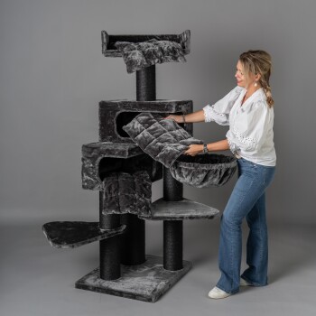 Eine Frau passt plüschige graue Kissen auf einem mehrstöckigen Katzenbaum an, der verschiedene Plattformen und Verstecke bietet, vor einem neutralen Hintergrund.