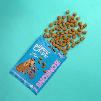 Un paquet de friandises pour animaux de compagnie Edgard Cooper, avec des chiens de dessin animé, déverse de petits cubes marron sur un fond turquoise.