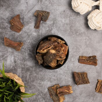 Eine schwarze Schüssel gefüllt mit verschiedenen Stückchen getrockneter Fleischleckerlis für Haustiere, umgeben von zusätzlichen Fleischstücken und einem geflochtenen Korb mit grünem Gras.