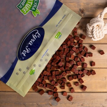 Eine Tüte mit Haustierleckereien im blau-gelben Design, die kleine, quadratische, rötlich-braune Stücke auf eine Holzoberfläche fallen lässt.