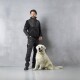 Eine Person in einer schwarzen Jacke und Hose steht neben einem lächelnden Golden Retriever vor einem grauen Hintergrund.