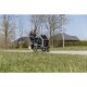 Eine Frau joggt mit einem Haustierwagen mit einer transparenten Kabine, im Park mit Bäumen und einem Haus im Hintergrund.