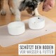 Ein Hund steht neben zwei weißen Schalen auf einer Matte; eine hat Wasser, die andere ist leer. Text: "SCHÜTZT DEN BODEN VOR WASSER & FUTTER."