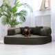 Ein schwarz-brauner Dackel, der auf einem plüschigen, olivgrünen Hundebett mit zwei kleinen Kissen entspannt, umgeben von einem hellen, luftigen Raum mit einer Palme.
