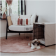 Ein kleiner, flauschiger Hund liegt auf einem rosa Teppich neben einer offenen Haustierbox in einem stilvollen Raum mit einem modernen Stuhl und dekorativen Kissen.