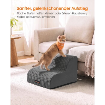 Rötliches Kätzchen mit orangefarbener Bandana klettert auf graue, strukturierte Haustiertreppen neben dem Sofa; sichtbarer deutscher Text 'Sanfter, gelenkschonender Aufstieg'.