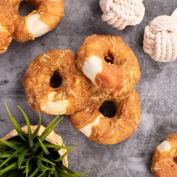 Drei Hundeleckerlis in Form von Bagels, aus einer goldbraunen, zähen Textur, umgeben von einer Kunstpflanze und Seilspielzeugen auf einer grauen Oberfläche.