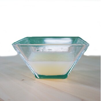 Eine klare Glasschüssel, gefüllt mit einer hellgelben Flüssigkeit, auf einer Holzoberfläche vor einem grünen Hintergrund.