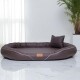 Braunes ovales Haustierbett mit Kissen und weißer Paspelierung, auf Marmorboden neben einer Pflanze und transparenten Vorhängen.