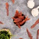 Eine schwarze Schüssel mit getrockneten Fleischleckerlis für Haustiere, umgeben von verstreuten Leckerlis, einem geflochtenen Korb mit Gras und zwei weißen Seilspielzeugen.