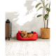 Ein flauschiger Pomeranian entspannt sich in einem rot-schwarzen Haustierbett inmitten stilvoller Dekoration, einschließlich eines Holzschranks, einer Pflanze und eines geflochtenen Korbs.