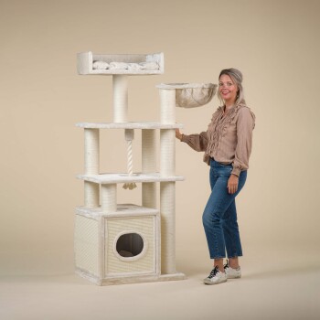 Eine Frau steht neben einem hohen, beigen Katzenbaum mit mehreren Ebenen, einer gemütlichen oberen Liegefläche, einer Hängematte und einem Kratzbaum.