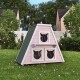 Ein pinkfarbener dreieckiger Katzenhaus mit drei Öffnungen, auf grünem Gras in einem Garten mit Bäumen und einem weißen Zaun im Hintergrund.