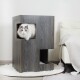 Eine flauschige weiße Katze mit blauen Augen, die aus einem modernen, dunklen Holz-Katzenhaus mit Ausschnitten hervorlugt, das neben einem weißen Sessel steht.