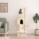 Eine getigerte Katze sitzt auf einem beigen Kratzbaum mit mehreren Ebenen und einem gemütlichen Versteck, neben einem grünen Sofa und einer Topfpflanze.