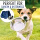 Ein Hund trägt eine Edelstahlschüssel, mit einer blauen Matte und einer weiteren Schüssel in der Nähe. Text: "PERFEKT FÜR DAHEIM & UNTERWEGS."