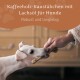 Ein weißer Hund mit einem Kaffeholz-Kaustäbchen im Mund, während eine Hand sich ausstreckt. Text: "Kaffeholz-Kaustäbchen mit Lachsöl für Hunde."