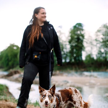 Eine Frau in einer schwarzen Jacke steht an einem Fluss mit einem braun-weißen Hund und zeigt eine natürliche Umgebung im Freien.