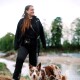 Eine Frau in einer schwarzen Jacke steht an einem Fluss mit einem braun-weißen Hund und zeigt eine natürliche Umgebung im Freien.