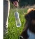 Eine Person hält eine tragbare Haustier-Wasserflasche mit einem silbernen Deckel und einem klaren Behälter, steht auf einem grasbewachsenen Feld mit einem Hund in der Nähe.