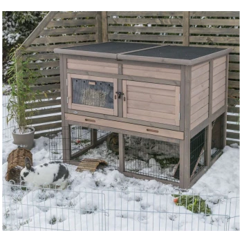 Ein Holzkaninchenstall mit einem schrägen Dach, das eine klare Tür zeigt, die Heu im Inneren zeigt, in einem schneebedeckten Außenbereich mit einem Kaninchen in der Nähe.
