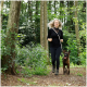 Eine Frau in schwarzer Sportkleidung joggt durch ein bewaldetes Gebiet mit einem braunen Hund, umgeben von grünem Laub und Bäumen.