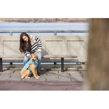Eine Frau in einem gestreiften Pullover sitzt auf einer Bank am See und streichelt sanft einen goldenen Hund, der einen grünen Mantel trägt.