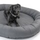 Ein schwarzer Labrador, der auf einem großen, runden, grauen Hundebett ruht und mit einem ruhigen Ausdruck direkt in die Kamera schaut.