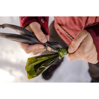 Eine Person hält eine Hundeleine mit einem "RUFF"-Logo, während sie auch einen gebundenen grünen Müllsack festhält und auf Schnee steht.