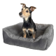 Ein kleiner, gemischter Hund mit einem schwarz-braunen Fell sitzt aufrecht in einem plüschigen, grauen Haustierbett mit einem weichen, pelzigen Rand.