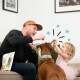Un enfant en robe rose tient une friandise tandis qu'un chien avec un collier bleu regarde avec impatience. Un sac de friandises pour animaux est sur le canapé.