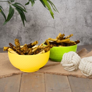 Zwei bunte Schalen gefüllt mit Hundeleckerlis, eine gelbe und eine grüne, auf einer Holzoberfläche mit einer Juteschicht platziert, neben zwei weißen Seilspielzeugen.