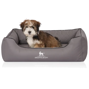 Ein kleiner, flauschiger Welpe sitzt bequem in einem grauen Hundebett mit einem gesteppten Design, mit dem Logo 'KNUFFELWUFF'.