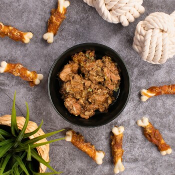 Eine schwarze Schüssel, gefüllt mit stückigem Haustierfutter, steht auf einer grauen Oberfläche, umgeben von hundeknochenförmigen Leckerlis und einem geflochtenen Korb mit grünem Gras.