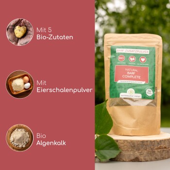 Ein Hund steht im Freien neben einem Sack "Natural BARF Complete" Ergänzungspulver, mit Dosierungsanweisungen auf Deutsch an der Seite.
