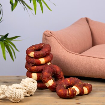 Ein Stapel von fünf roten und weißen Hundekaukringern liegt auf einem Holztisch neben einem weißen Seilspielzeug, mit einem rosa Haustierbett im Hintergrund.