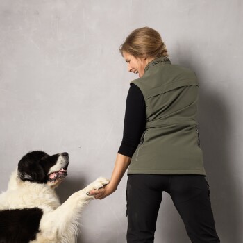 Eine Frau in einer grünen Weste interagiert mit einem großen, flauschigen schwarz-weißen Hund, der seine Pfote hebt.